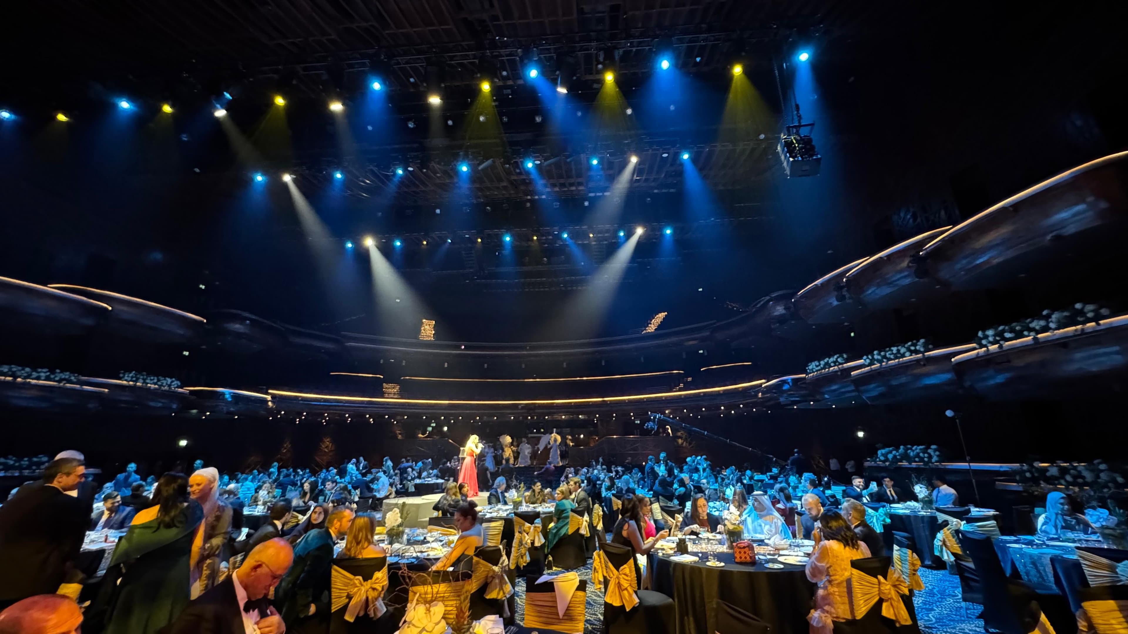 Formal banquet in a grand theater with guests seated under dramatic blue and yellow spotlights.